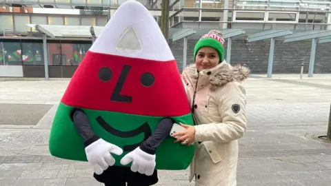 Urdd Mah in a large cream coloured coat and green & white beanie posing with the Urdd's Mascot - which is a triangle with red, white and green stripes and face on it in Cardiff Bay - outside the Senedd.