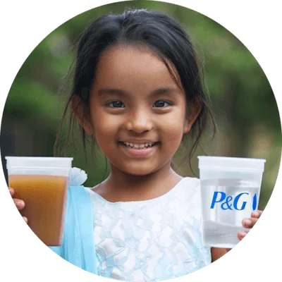  A small, young Indian girl in a blue shirt holding two cups. The cup in her left hand has dirty water and the cup in her right hand has clean water, with a blue text logo that says “P&G.”